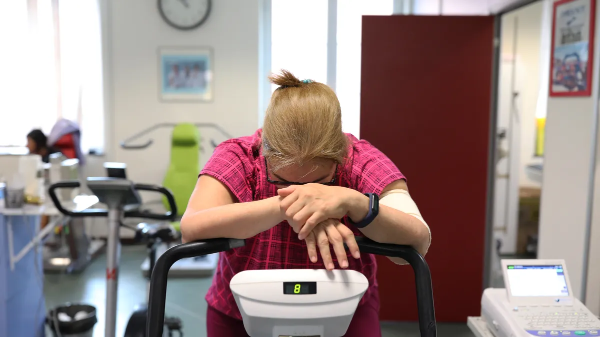 A woman with brown hair clipped up, wearing a pink shirt, bends forward on an exercise bike at a hospital, exhausted.