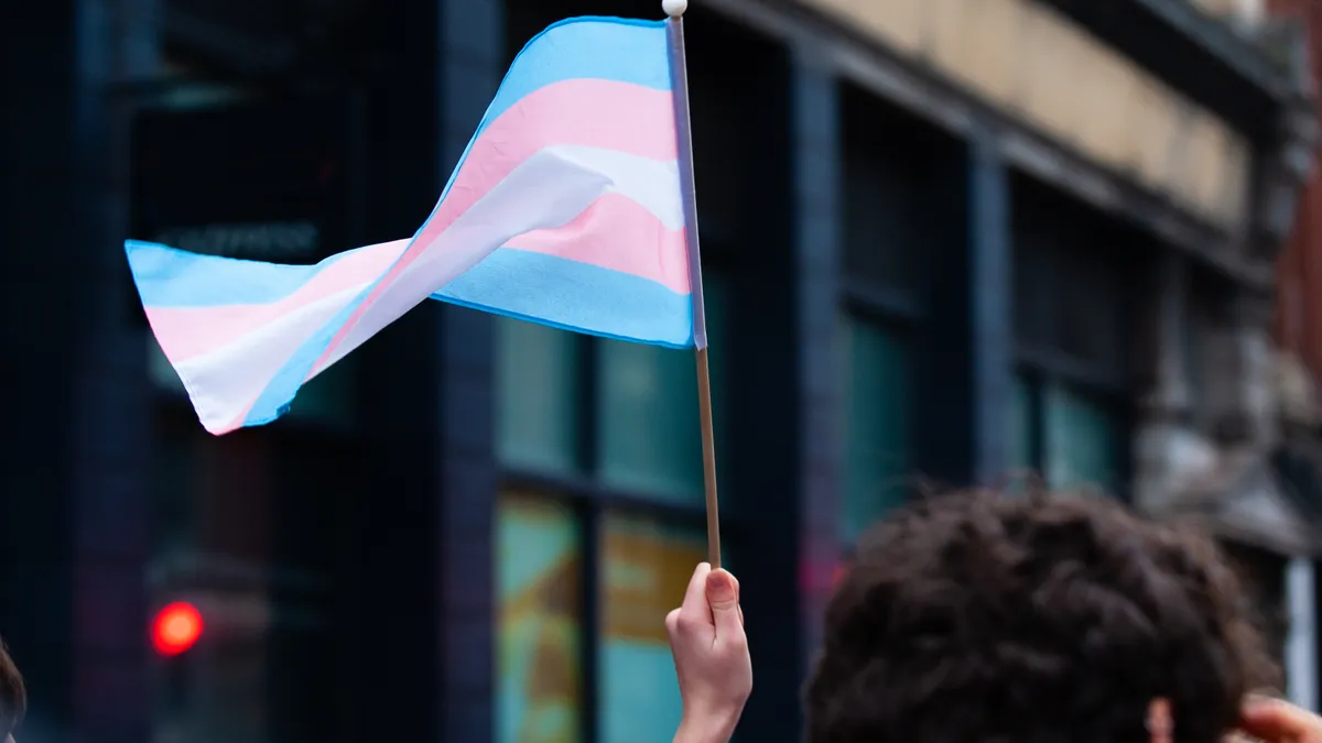 A hand holds up a small transgender pride flag in a Bristol Street. The transgender pride flag is a symbol of pride for the trans community. The blue and pink stripes represent traditional colours for