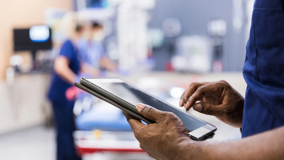 A healthcare worker in blue scrubs holds a tablet in a clinical setting