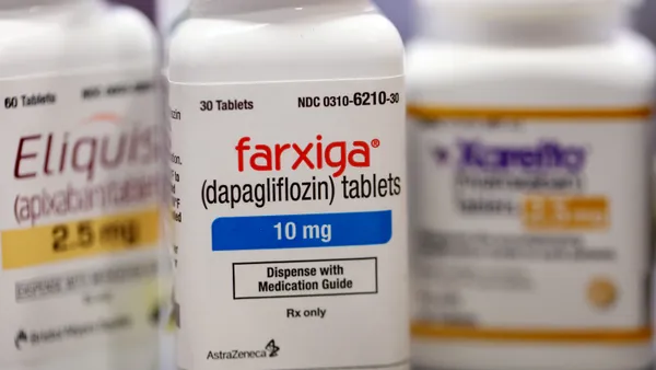 Pills bottles labeled with the logos for Eliquis, Farxiga and Xarelto are arranged on a table.