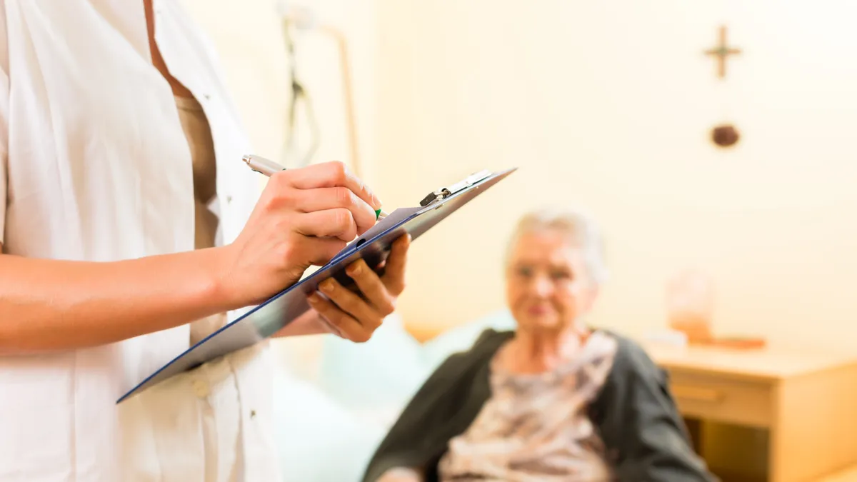 An elderly woman sits in a doctor's office as a medical worker writes on a clipboard.